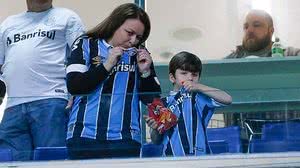 Taís Dias e o filho Bernardo beijam o escudo do Grêmio antes de jogo no dia 25 | Folhapress