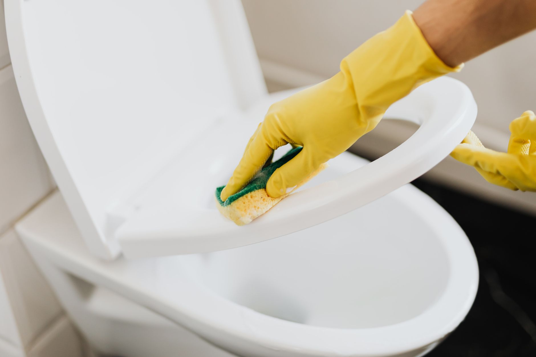 Uma pessoa usando luvas amarelas e uma esponja para limpar um assento de vaso sanitário dentro de casa.