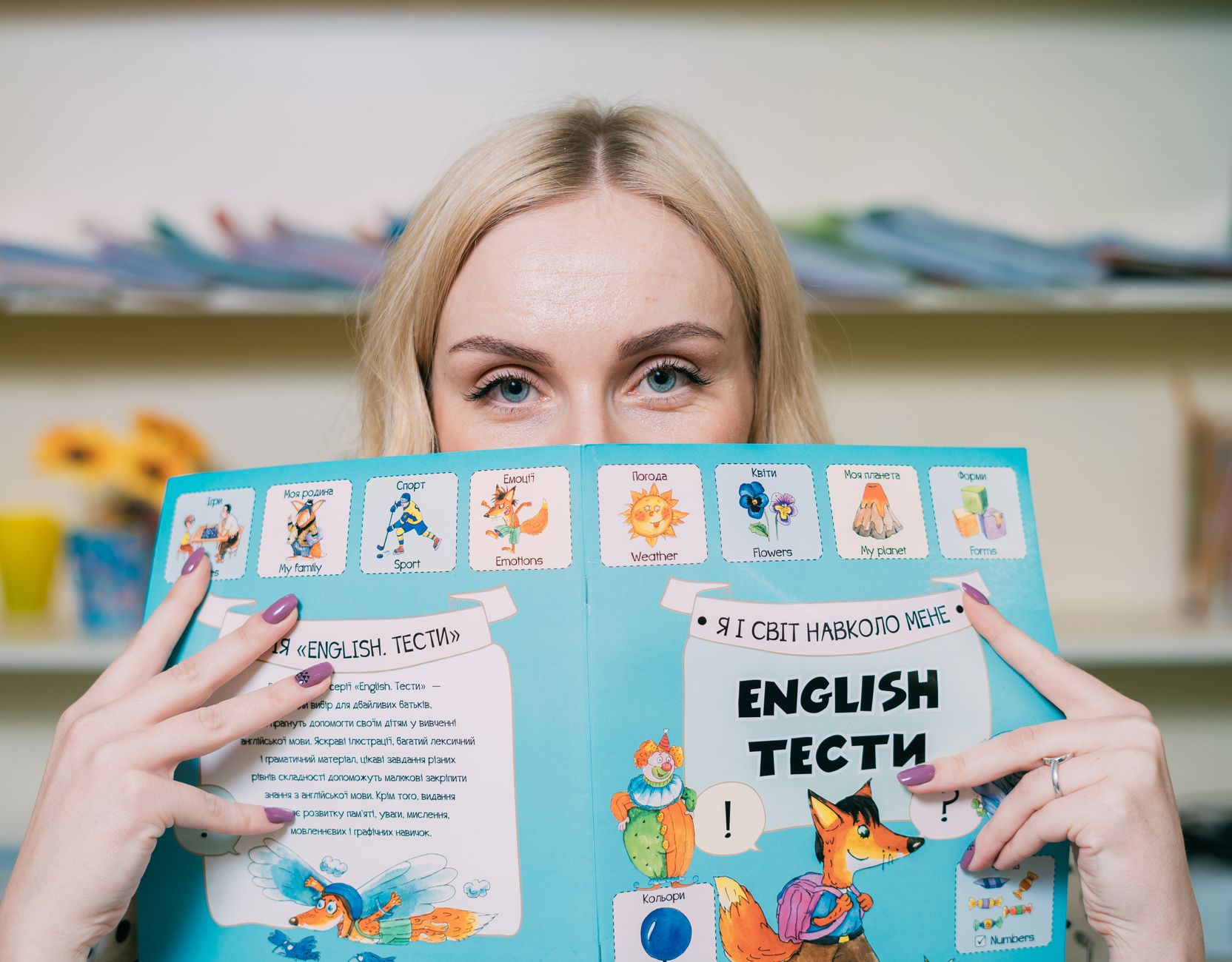 Mulher loira segurando e lendo um livro ilustrado em inglês dentro de casa.