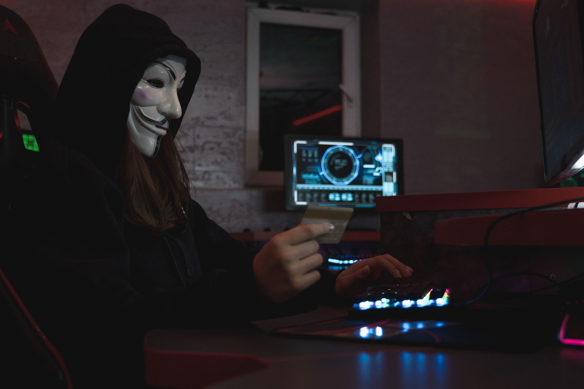 Hacker mascarado com cartão de crédito no computador, simbolizando cibercrime e anonimato.