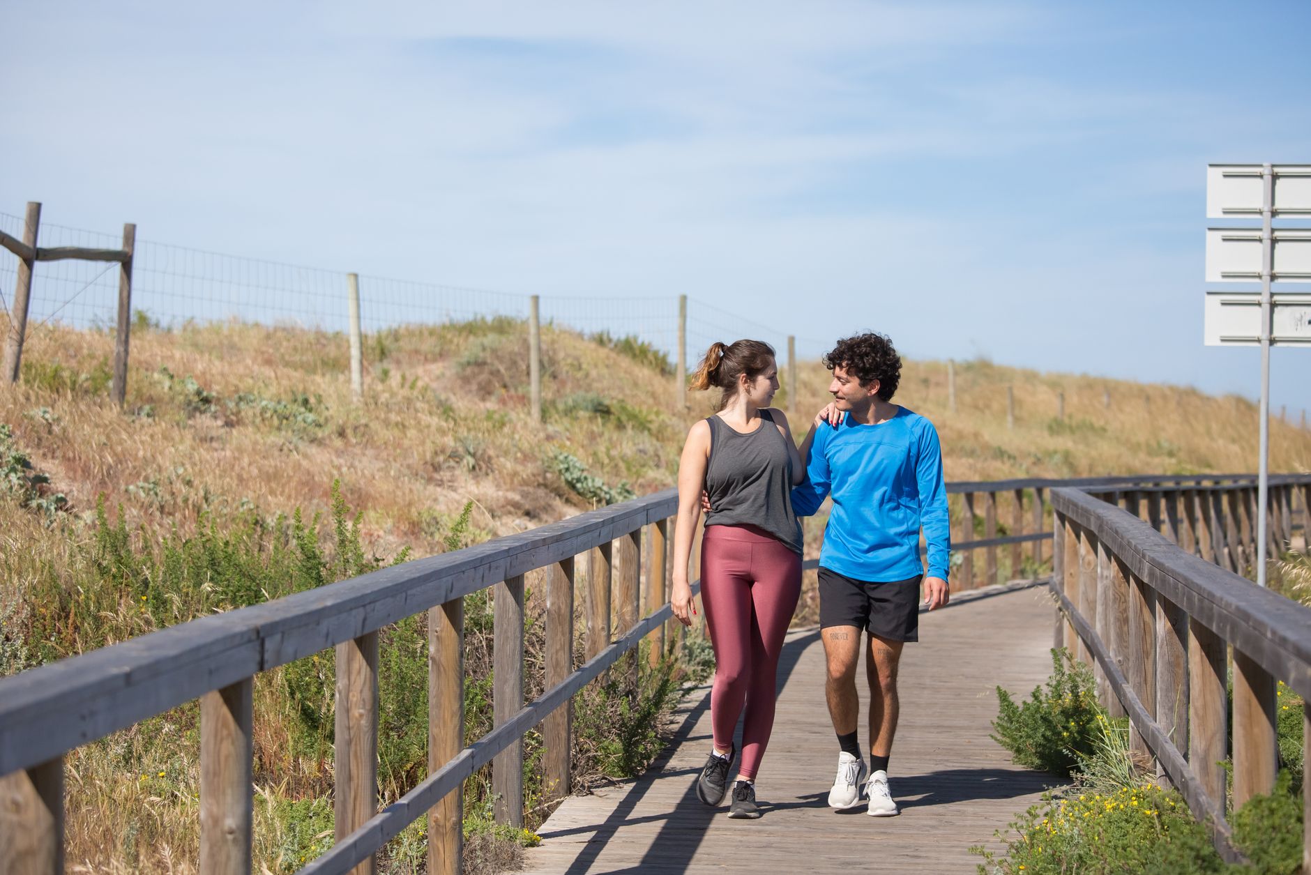 Um casal vestido com roupas esportivas passeando juntos em um calçadão ensolarado em um ambiente natural.