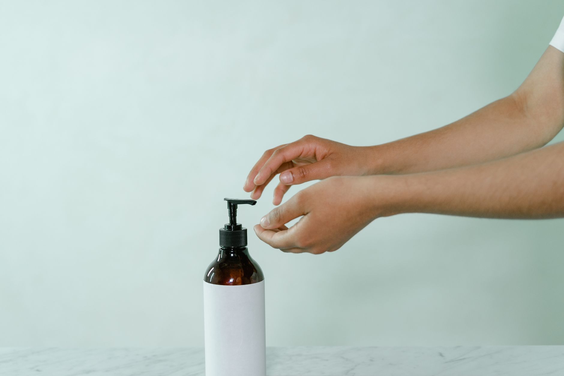Uma foto minimalista de mãos alcançando um frasco de bombeamento com um rótulo em branco em um fundo branco.