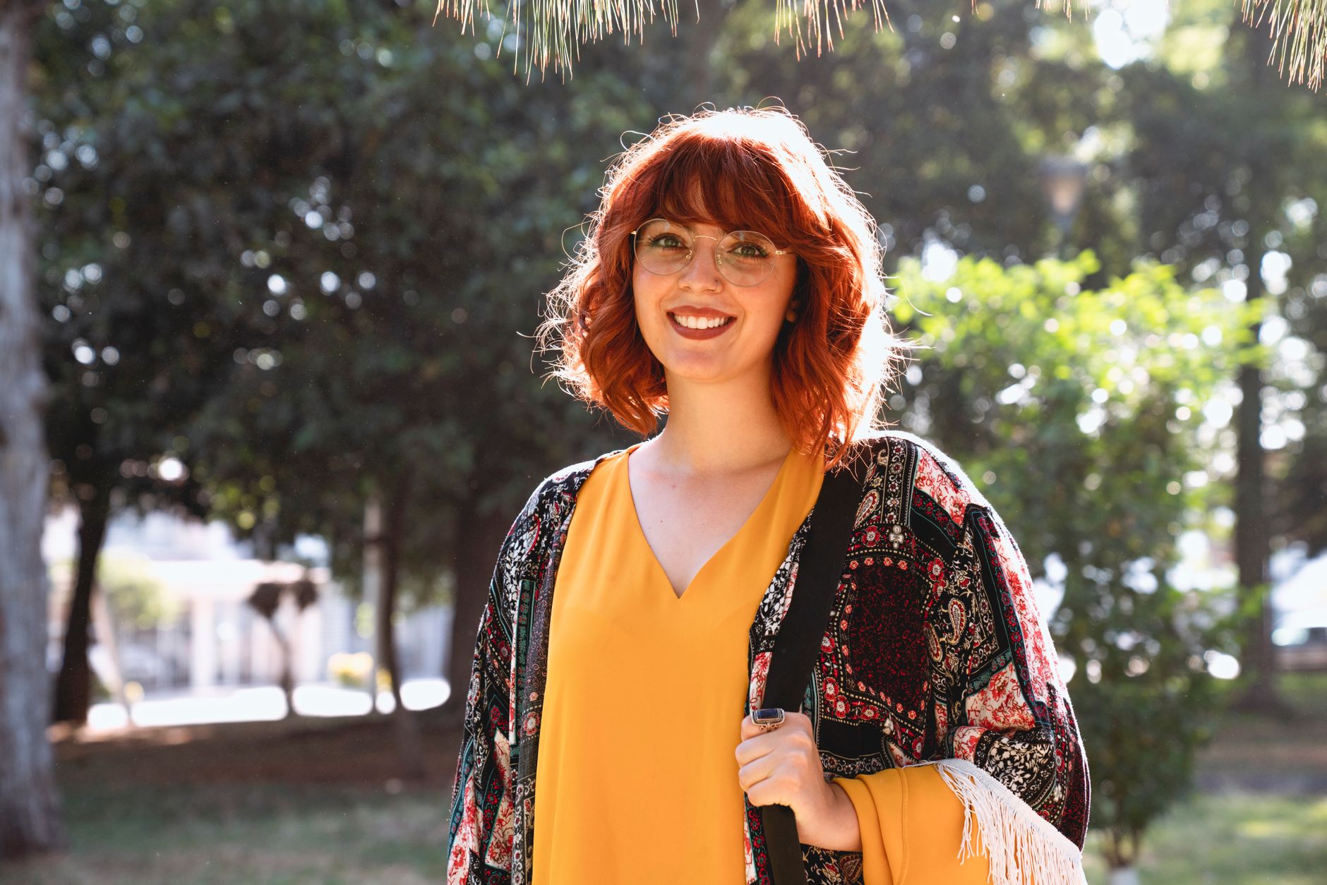 Mulher ruiva sorridente com óculos desfrutando de um dia ensolarado ao ar livre, usando um xale colorido.