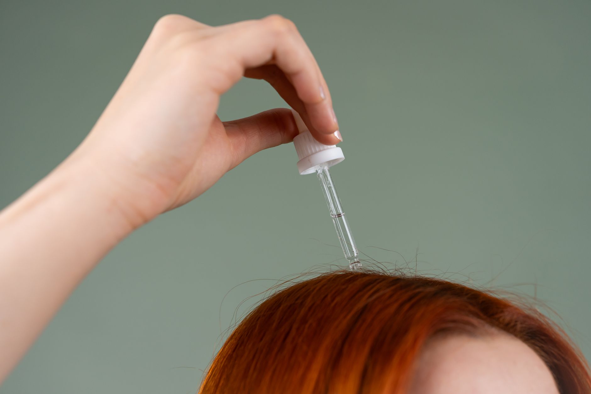 Close-up de uma mão aplicando sérum capilar em cabelo vermelho com um conta-gotas.